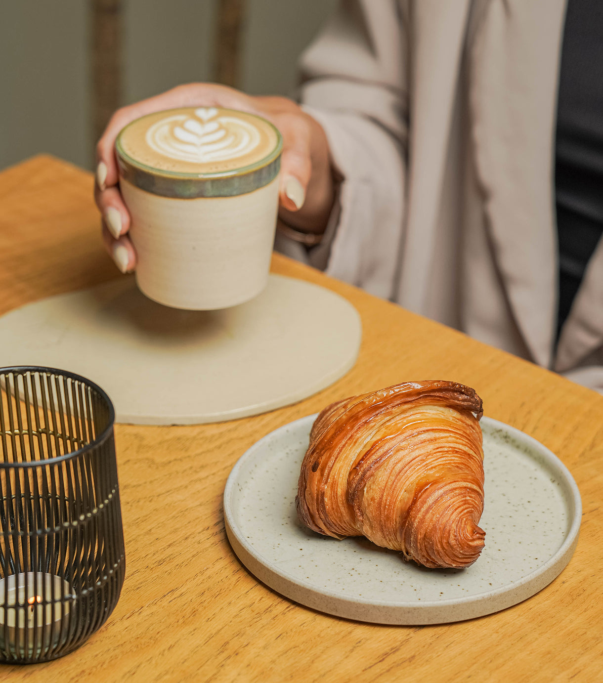 Person holding a Matcha cup next to a croissant on a wooden table in Dubai