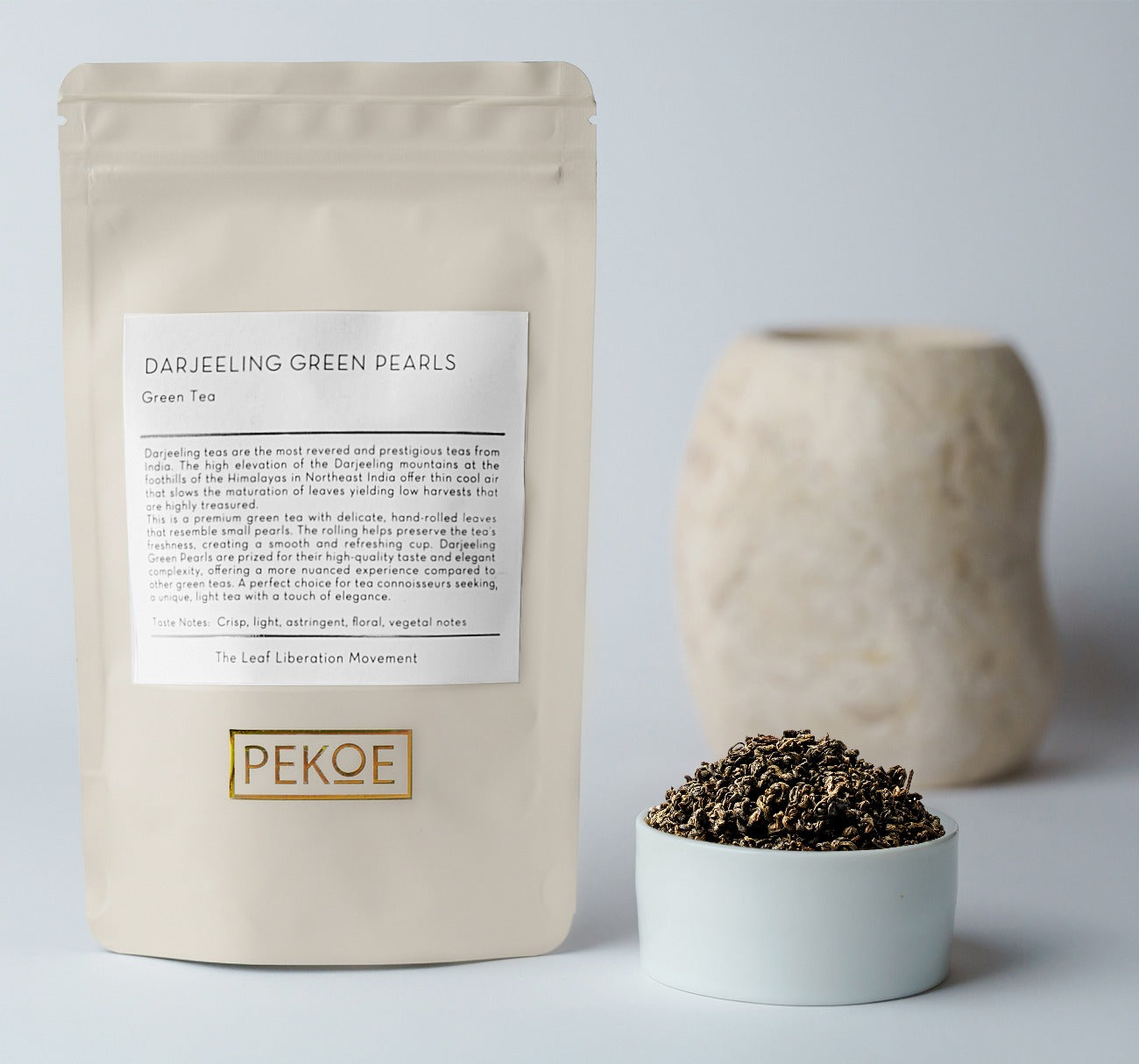 A stand-up pouch of Darjeeling Green Pearls tea by Pekoe, shown next to a small white bowl filled with the loose tea leaves and a blurred ceramic vase on a light gray background.
