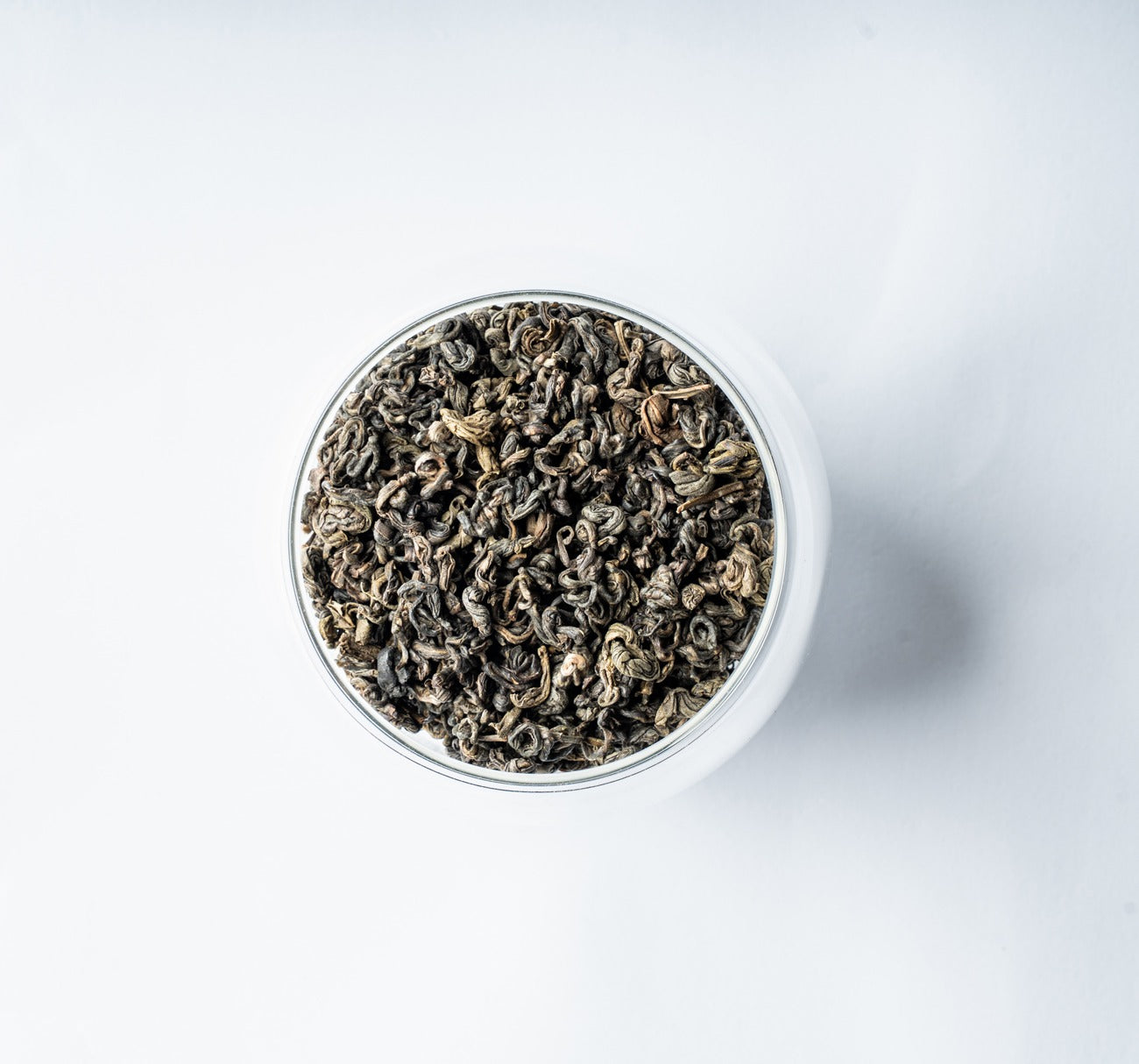 A top-down view of dark, curly loose-leaf Darjeeling Green Pearls tea filling a clear glass jar, set against a clean white background.