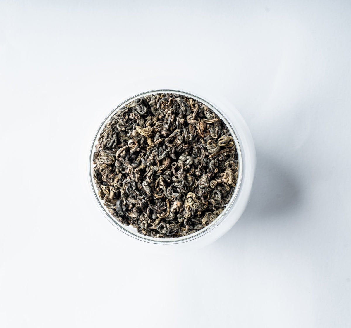 A top-down view of dark, curly loose-leaf Darjeeling Green Pearls tea filling a clear glass jar, set against a clean white background.